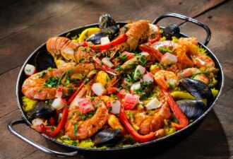 Плато з паельї з морепродуктами, включає креветки, мідії, шматочки крабового м'яса, приправлене свіжою зеленню та перцем на яскравому рисовому фоні.