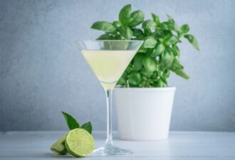 Коктейль у елегантному келиху з лимонним відтінком на фоні зеленого базиліку в горщику та скибочок лайма з листям м'яти.