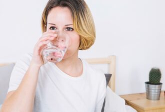 Жінка у світлому одязі п'є воду з прозорого стакана, сидячи на ліжку в світлій кімнаті. На столі поруч знаходиться кактус у горщику.
