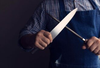 Чоловік у фартусі тримає кухонний ніж і точило, демонструючи процес заточування ножів.