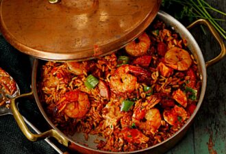 Каструля з рисом, креветками та овочами, покрита мідною кришкою, на фоні текстурованого столу. Страва виглядає апетитно, з яскравими інгредієнтами та привабливою подачею.