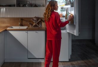 Жінка у червоному піжамі стоїть біля відкритого холодильника в сучасній кухні, вибираючи напій в затишній обстановці.