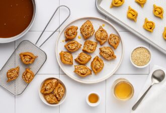Марокканське печиво шебакія, вкрите медом і кунжутом, на білому тарілці, оточене сирими заготовками, мискою з кунжутом, ложкою, ситом і каструлею з медовим сиропом на світлій плитці.