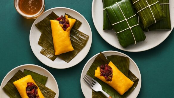 Гаяки з яскраво-жовтого кукурудзяного тіста, з м’ясною начинкою, розкладені на керамічних тарілках поверх пофарбованого бетонного столу. Страва загорнута в бананове листя, вид зверху, яскраве студійне освітлення.