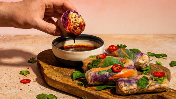 Літні роли з куркою, овочами та хойсін-соусом на дерев’яній дошці, поруч листя кінзи й перцю чилі, з чоловічою рукою, що тримає рол над соусом.
