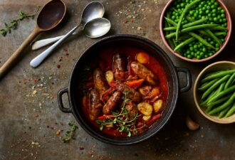 Домашній хот-пот із ковбасками, перцем та картоплею в чавунному казані на потертій дерев’яній поверхні, поруч зелена квасоля й горох, розсипані крихти та приправи.