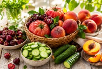 Кошик із сезонними літніми продуктами на дерев’яному столі: стиглі рожеві томати, соковиті персики, темна черешня та свіжі огірки. Композицію доповнюють зелень і м’яке сонячне світло, що створює атмосферу початку літа та щедрого врожаю.