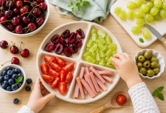 Вид зверху на дитячу тарілку з безпечно підготовленими продуктами для перекусу. Черешня без кісточок розрізана вздовж, виноград нарізаний поздовжніми частинами, помідори чері поділені на четвертинки, а сосиски нарізані довгими смужками. Поруч розташовані миски з ягодами та овочами, а дитина тягнеться до тарілки. Фото ілюструє правила безпечної подачі їжі для маленьких дітей та профілактику задухи під час прийому їжі.